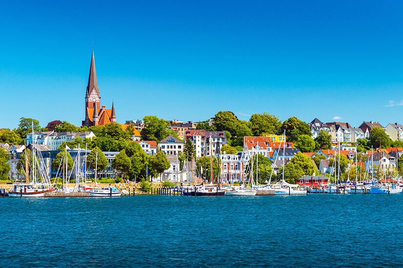 Segelboote und Yachten liegen vor der lebendigen Stadtkulisse des Flensburger Hafens. Über den bunten Dächern erhebt sich der markante Turm der St.-Jürgen-Kirche. Tiefblaues Wasser, historische Fassaden und maritime Details prägen das nordische Panorama. Hundestrände Ostsee Flensburg