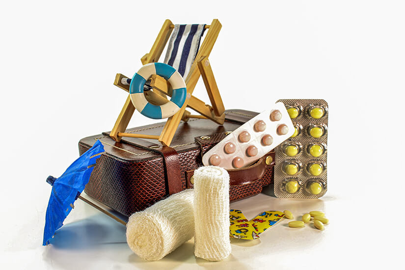 Auf einem braunen Miniaturkoffer mit Liegestuhl, Rettungsring und Cocktailschirmchen sind verschiedene Medikamente in Blistern, Mullbinden, bunte Kinderpflaster und einzelne Tabletten arrangiert. Reiseapotheke Basis