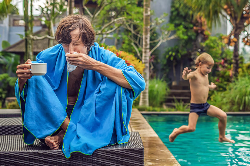 Kränklich in ein blaues Handtuch gehüllt sitzt ein Mann am Beckenrand, hält eine Tasse Tee in der Hand und putzt sich die Nase, während im Hintergrund sein Sohn fröhlich in den Pool springt. Reiseapotheke Vater Sohn