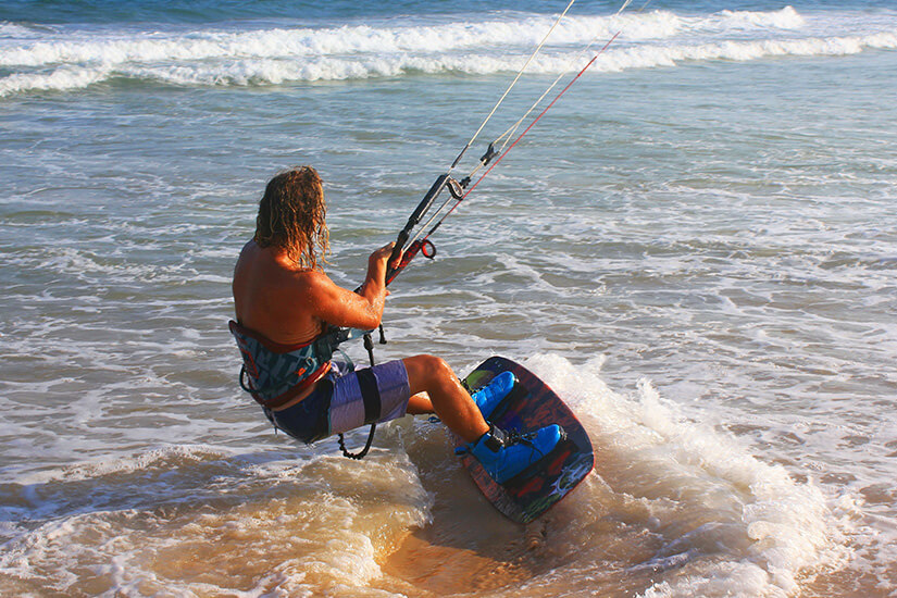 Ein Mann mit schulterlangen Haaren steht auf einem Board im seichten Meerwasser. Die Schnüre eines Lenkdrachen sind zu sehen, der ihn ins offene Wasser zieht. Kitesurfing Teaser