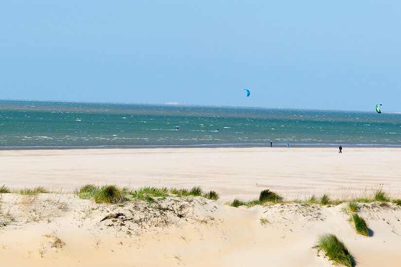 Ein langer heller Sandstrand ist zu sehen. Über dem Wasser schwebt ein blauer Lenkdrachen. Kitesurfing Brouwersdam