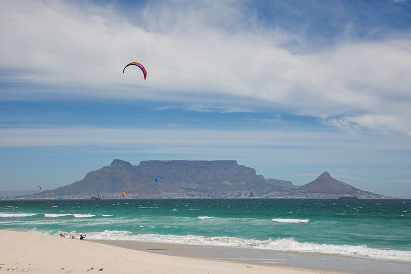 Lenkdrachen schweben über türkisfarbenem Meer vor der Küste Kapstadts. Im Hintergrund ist der Blouberg zu sehen. Kitesurfing Kapstadt