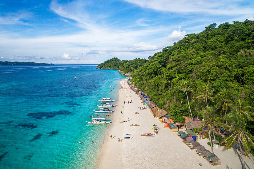 Ein langer heller Sandstrand liegt zwischen tropischer Vegetation und klarem Meer in verschiedenen Blautönen. Schiffe treiben im Wasser, am Strand tummeln sich Menschen mit Surfbrettern. Kitesurfing Boracay