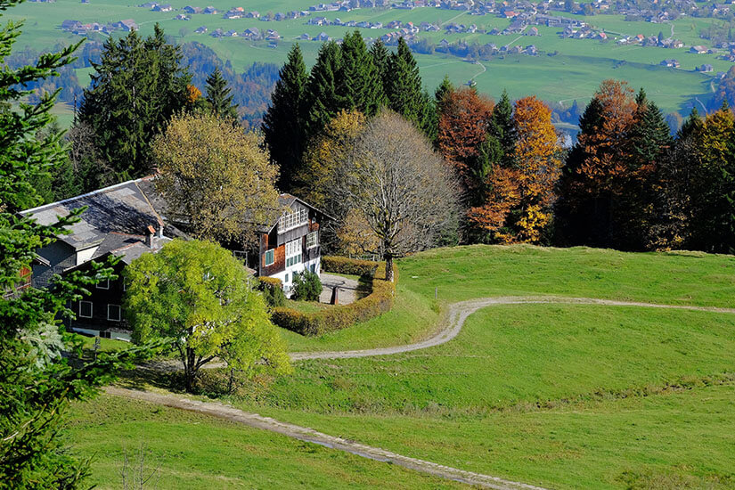 Berghaus am Bödele oberhalb von Dornbirn im Herbst. Bunte Laubbäume und Wiesen umgeben das Gebäude. Im Tal sind vereinzelte Siedlungen sichtbar. Dornbirn Österreich Bödele