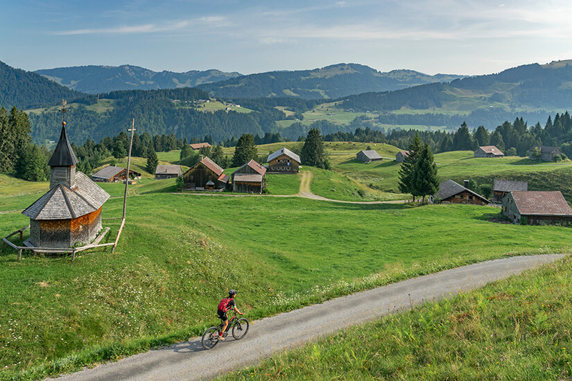 Ein Radfahrer ist auf einer schmalen Straße zwischen grünen Wiesen und traditionellen Holzhäusern unterwegs. Im Vordergrund steht eine kleine Kapelle, dahinter breitet sich eine hügelige Landschaft aus. Dornbirn Österreich Fahrrad