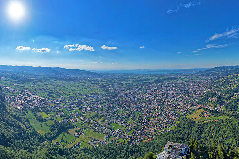 Weite Aussicht über Dornbirn und das Rheintal bei klarem Wetter. Im Hintergrund ist der Bodensee zu erkennen, während sich Wohngebiete, Felder und bewaldete Hänge über die Landschaft verteilen. Dornbirn Österreich Aussicht