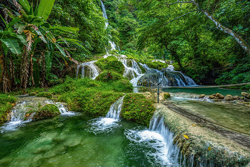 Eine terrassenförmige Wasserfalllandschaft der Mele Cascades inmitten eines dichten, tropischen Regenwaldes. Klares, grünlich schimmerndes Wasser fließt über moosbedeckte Steinstufen und bildet mehrere kleine, natürliche Becken. Im Vordergrund erstreckt sich ein flacher Pfad mit einem einfachen Seilgeländer zur Sicherung, während im linken Hintergrund eine große Bananenstaude mit breiten Blättern aufragt und im fernen Hintergrund ein höherer Wasserfall durch das dichte Blätterdach blickt. Vanuatu Mele Cascades