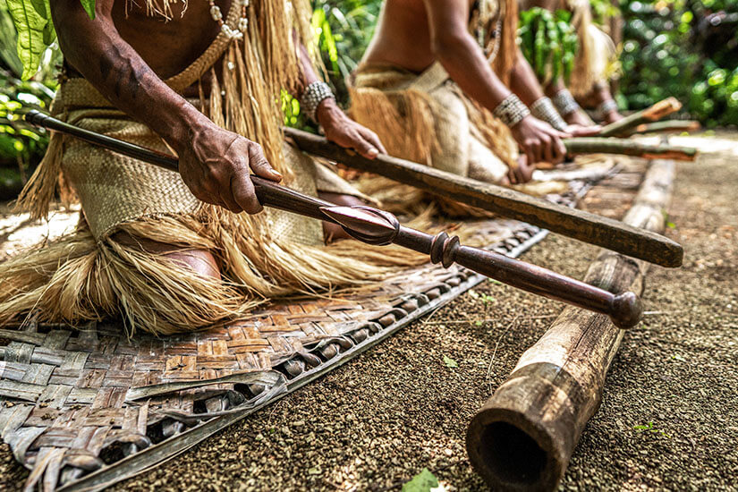 Eine detailreiche Nahaufnahme von mehreren Männern auf Vanuatu, die in traditioneller Kleidung in einer Reihe auf einer großen, geflochtenen Matte aus Naturfasern knien. Sie tragen traditionelle Röcke aus hellen Pflanzenfasern und gemusterte Armbänder an den Handgelenken. In den Händen halten sie lange, glatt polierte Holzstäbe, mit denen sie rhythmisch auf ein dickes, hohles Bambusrohr schlagen, das vor ihnen auf dem sandigen Boden liegt. Der Fokus liegt scharf auf den Händen und dem kunstvoll geschnitzten Stab im Vordergrund, während die im Hintergrund Knienden und die grüne Vegetation weich gezeichnet sind. Vanuatu Braeuche