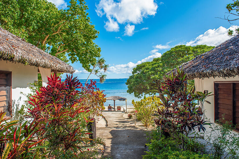 Ein befestigter Weg führt zwischen zwei traditionellen, weiß getünchten Bungalows mit strohgedeckten Dächern hindurch direkt zum Strand. Im Vordergrund säumen prächtige, rot-grün gemusterte tropische Sträucher den Pfad. Am Ende des Weges öffnet sich der Blick auf den feinen Sandstrand, das tiefblaue Meer und ein kleines, strohgedecktes Sonnendach mit zwei Liegestühlen unter einem strahlend blauen Himmel mit weichen, weißen Wolken. Vanuatu Strandblick