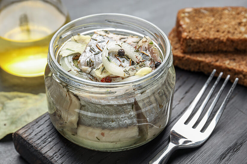 Zu erkennen ist ein Glas mit eingelegtem Hering, garniert mit Zwiebelringen, Kräutern und Gewürzen in einer Marinade. Daneben liegen Scheiben dunkles Brot, eine Gabel und im Hintergrund eine Flasche mit hellem Öl oder Essig. Estland Essen Hering