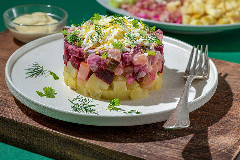Zu erkennen ist ein geschichteter Salat aus gewürfelten Kartoffeln, Roter Bete, Fisch und weiteren Zutaten, garniert mit geriebenem Ei sowie frischen Kräutern. Daneben stehen eine Gabel und eine kleine Schale mit heller Sauce, während im Hintergrund weitere vorbereitete Zutaten zu sehen sind. Estnisches Essen Rosolje