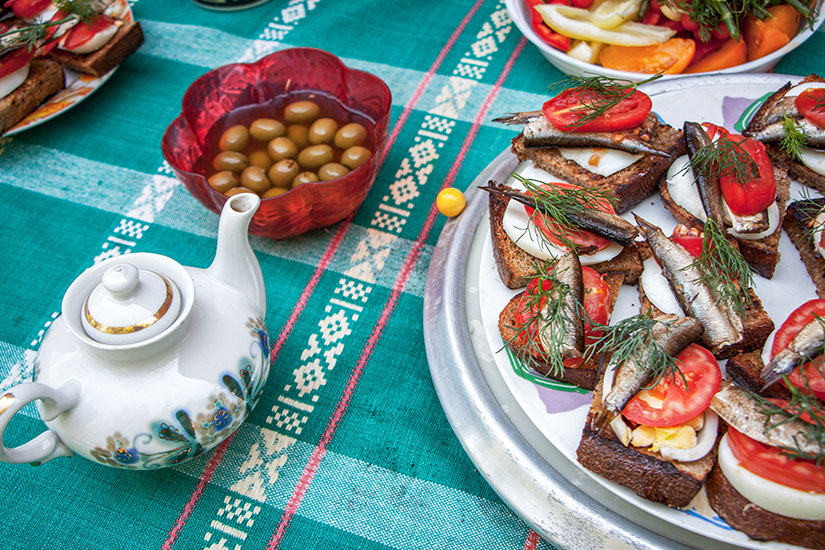 Zu erkennen ist eine Auswahl belegter Brote mit kleinen Fischen, Tomatenscheiben, Ei und frischem Dill auf einer Servierplatte. Daneben stehen eine Schale mit grünen Oliven, eine dekorative Teekanne sowie weitere Speisen auf einem türkisfarbenen Tisch. Estland Essen Kiluvoileib