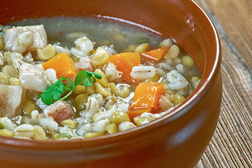 Zu erkennen ist eine herzhafte Suppe mit Erbsen, Gerste oder Graupen, Karottenwürfeln und verschiedenen Fleischstücken in einer braunen Schüssel. Als Garnitur dient ein kleines frisches Blattgrün, während die Schüssel auf einem rustikalen Holztisch steht. Estland Essen Erbsensuppe