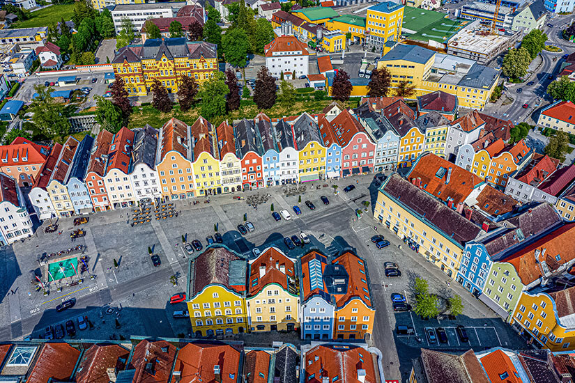 Zu erkennen ist eine historische Altstadt mit einem großen Marktplatz, der von farbenfrohen Bürgerhäusern mit giebelständigen Fassaden umgeben ist. Auf dem Platz stehen geparkte Autos und Außengastronomie, während sich im Hintergrund weitere Gebäude und Grünflächen erstrecken. Schaerding Stadt