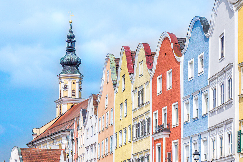 Zu erkennen ist eine Reihe farbenfroher historischer Bürgerhäuser mit geschwungenen Giebeln entlang einer Straße. Im Hintergrund erhebt sich der Turm einer Kirche mit markanter Zwiebelhaube vor einem bewölkten Himmel. Schaerding Silberzeile