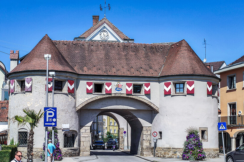 Zu erkennen ist ein historisches Stadttor mit zwei runden Türmen und einem großen Torbogen, durch den eine Straße in die Altstadt führt. Vor dem Gebäude befinden sich Straßenschilder, Blumenschmuck und einige Passanten, während über dem Tor eine Uhr und ein Wappen angebracht sind. Schaerding Stadttor