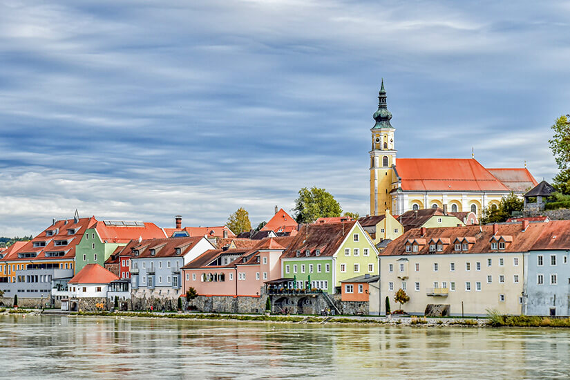 Zu erkennen ist eine historische Uferstadt mit pastellfarbenen Häusern entlang eines Flusses und einer erhöht gelegenen Kirche mit hohem Turm. Darüber ziehen dichte Wolken über den Himmel und spiegeln sich teilweise im ruhigen Wasser. Schaerding Kirche