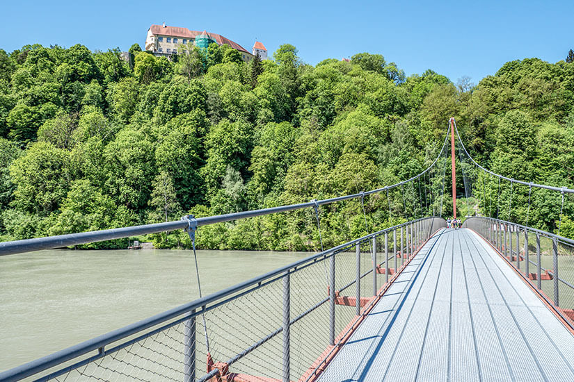 Zu erkennen ist eine schmale Hängebrücke, die über einen breiten Fluss zu einem dicht bewaldeten Hang führt. Auf der Anhöhe steht ein historisches Gebäude oder Schloss, während der Himmel klar und blau ist. Oesterreich Mariensteg