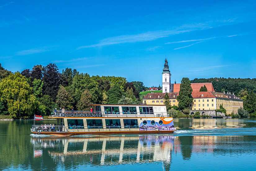 Zu erkennen ist ein modernes Ausflugsschiff, das über einen ruhigen Fluss fährt und sich im Wasser spiegelt. Im Hintergrund liegen ein Kloster oder eine Kirche mit hohem Turm sowie weitere historische Gebäude, umgeben von üppigen Bäumen unter einem klaren blauen Himmel. Inn Schifffahrt