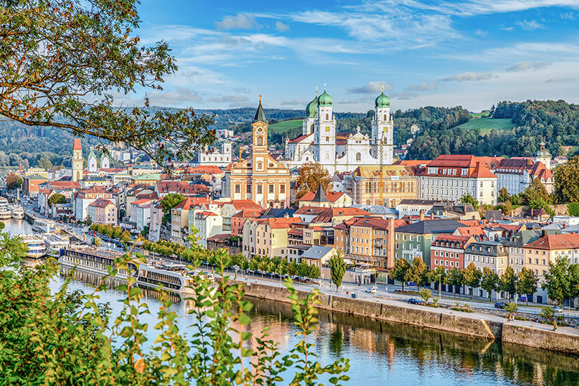 Zu erkennen ist eine historische Stadt am Fluss mit einer Uferpromenade, farbenfrohen Gebäuden und mehreren markanten Kirchen mit Türmen und grünen Kuppeln. Im Vordergrund rahmen Bäume die Aussicht ein, während sich hinter der Altstadt sanfte Hügel und bewaldete Landschaften erstrecken. Bayern Passau