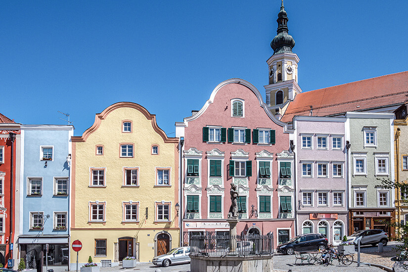 Zu erkennen ist eine Reihe pastellfarbener historischer Bürgerhäuser mit geschwungenen Giebeln an einem gepflasterten Platz. Im Hintergrund ragt der Turm einer Kirche mit Zwiebelhaube empor, während im Vordergrund ein Brunnen, geparkte Autos und Fahrräder zu sehen sind. Schaerding Altstadt