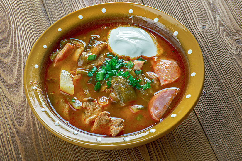 Zu erkennen ist eine kräftige Suppe mit Fleischstücken, Wurstscheiben, Kartoffeln, Gurken und weiteren Gemüseeinlagen in einer rötlichen Brühe. Obenauf befinden sich ein Klecks Sauerrahm und gehackte Frühlingszwiebeln, serviert in einer rustikalen Schüssel auf einem Holztisch. Estland Essen Seljanka