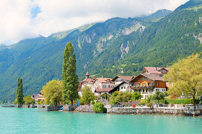 Das idyllische Dorf Brienz vom türkisblauen See aus betrachtet. Grüne Bäume und bergiges Hinterland prägen die Szene. Brienz Schweiz Wetter