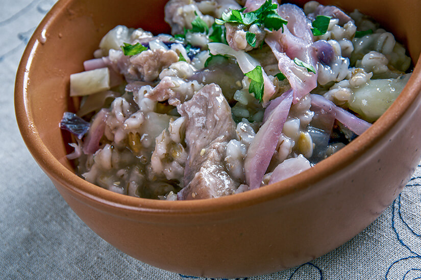 Zu erkennen ist eine herzhafte Speise mit Fleischstücken, Graupen und Sauerkraut oder Kohl in einer braunen Tonschüssel. Garniert wird das Gericht mit roten Zwiebelstreifen und frischer Petersilie. Estland Essen Mulgikapsad