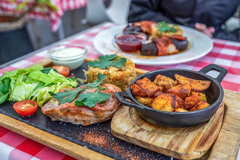 Zu erkennen ist ein rustikal angerichtetes Gericht mit einem gebratenen Schweinesteak, knusprigen Kartoffelspalten, Salat und einer Portion Sauerkraut. Im Hintergrund steht ein weiterer Teller mit Würsten und Dips auf einem Tisch mit rot-weiß karierter Tischdecke. Estland Essen Seapraad