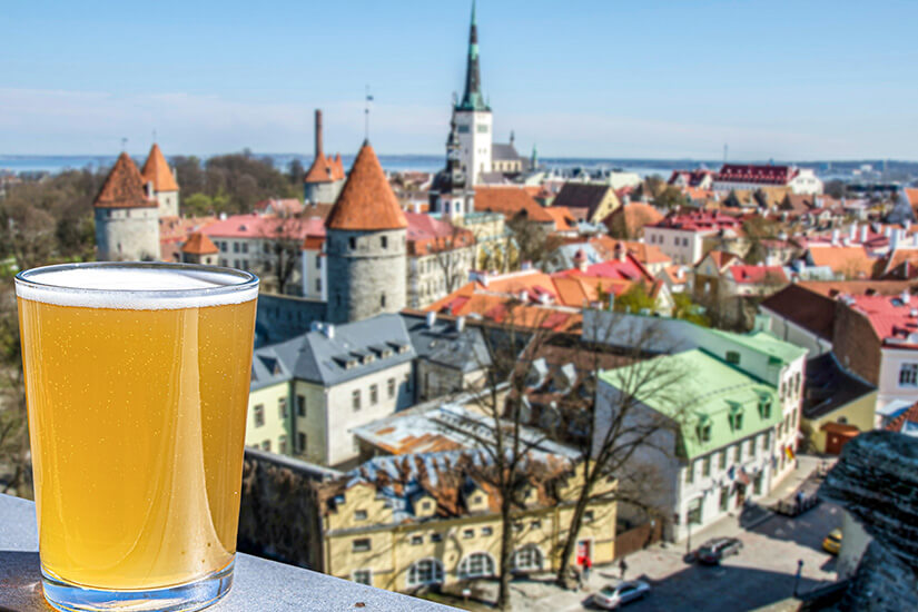 Zu erkennen ist ein Glas helles Bier auf einer Mauer oder Brüstung mit Blick über eine historische Altstadt. Im Hintergrund stehen mittelalterliche Türme, eine hohe Kirchturmspitze und zahlreiche Gebäude mit roten Dächern. Estland Trinken Bier