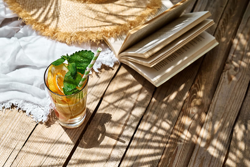Verlockend arrangiert stehen ein erfrischendes Glas Zitronenlimonade mit Minzblättern und Papierstrohhalm, ein aufgeschlagenes Buch und ein Strohhut auf einem sonnenbeschienenen Holzboden, während Palmenblätter Schattenmuster werfen. Urlaubslektuere Kolumbien