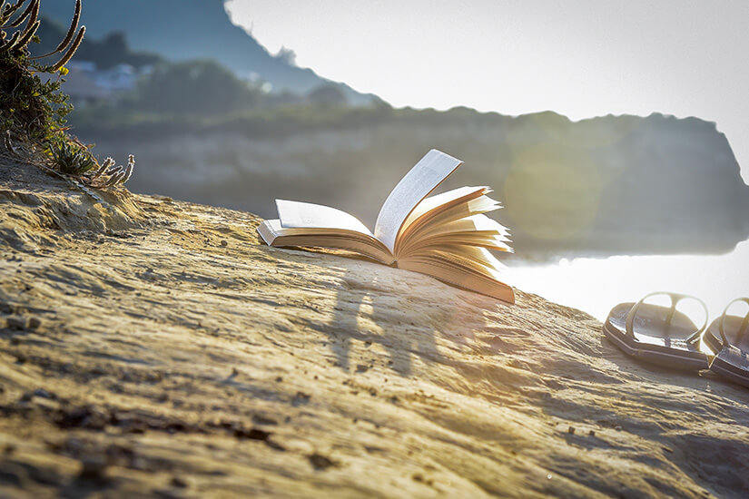 Sanft blättert der Wind in einem aufgeschlagenen Buch, das neben Flip-Flops auf einem sonnendurchfluteten Felsen an einer mediterranen Küste liegt. Urlaubslektuere Griechenland