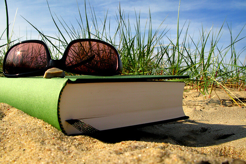 Halb im Sand vergraben liegt ein grünes Buch mit einer darauf abgelegten Sonnenbrille, umgeben von Strandhafer und unter strahlend blauem Himmel. Urlaubslektuere Ostsee