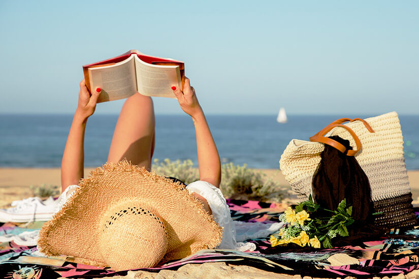 Entspannt auf einer bunten Decke liegend hält eine Frau mit Sonnenhut ein aufgeschlagenes Buch über sich, während neben ihr eine Strohtasche mit gelben Blumen steht und im Hintergrund das Meer mit einem Segelboot zu sehen ist. Urlaubslektuere Barcelona