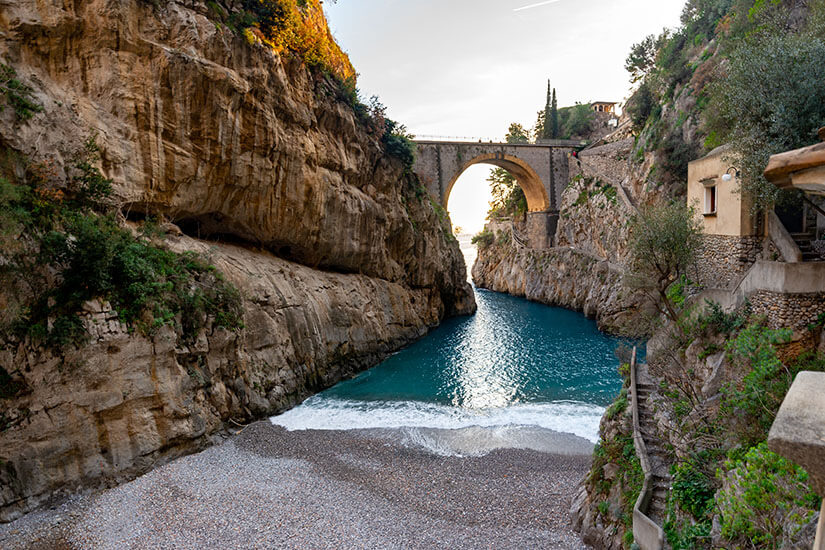 Zwischen zwei mächtigen Felswänden schimmert türkisblaues Wasser am Fiordo di Furore – einer geologischen Rarität an der Amalfiküste. Die imposante Steinbrücke überspannt die enge Schlucht und rahmt den Blick aufs offene Meer. Am Ende des Einschnitts liegt ein kleiner Kiesstrand, der zu den verstecktesten Badestellen der gesamten Costiera gehört. Amalfiküste Italien Fiordo di Furore