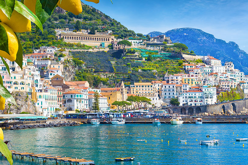 Pastellfarbene Häuser staffeln sich am Hang über dem Hafen von Amalfi, während im Vordergrund goldgelbe Zitronen ins Bild ragen. Die Szene vereint alles, wofür die Region berühmt ist: südländische Architektur, mediterranes Flair und die allgegenwärtigen Zitrusfrüchte. Fischerboote und kleine Jachten dümpeln im ruhigen Hafenbecken vor der malerischen Kulisse. Amalfikueste Italien Kampanien