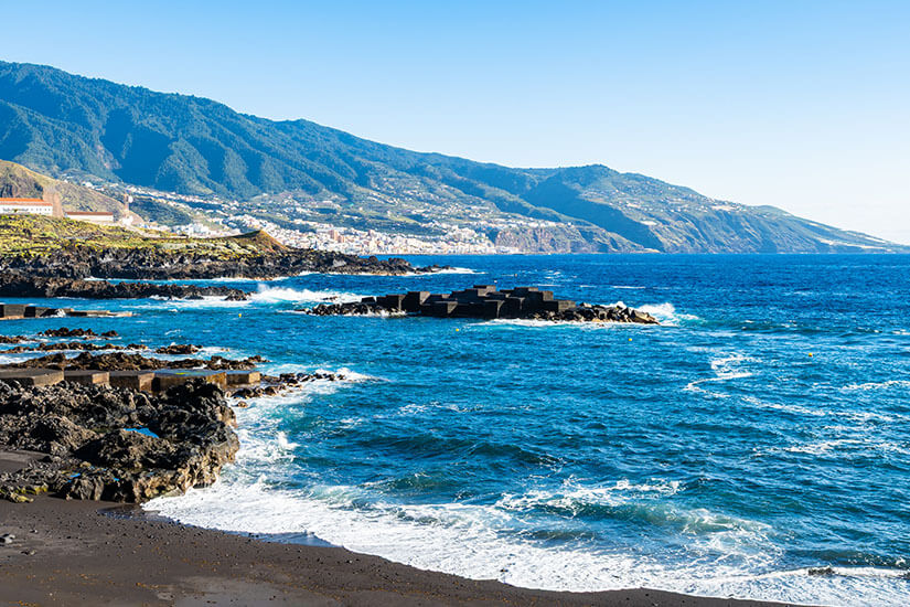 Ein Strandabschnitt der Playa de los Cancajos mit dunklem, vulkanischem Sand im Vordergrund. Die Wellen des kräftig blauen Atlantiks spülen weiße Gischt an die Küste. Dunkle Lavafelsen und künstliche Wellenbrecher aus Betonblöcken liegen vor dem Strand im Wasser. Im Hintergrund erstreckt sich eine Steilküste mit grünen, dicht bewachsenen Bergzugängen und einer kleinen Stadt an den Hängen unter einem klaren, blauen Himmel. La Palma Palma Spanien Playa de los Cancajos