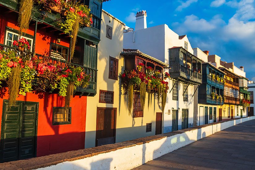 Eine historische Häuserzeile in Santa Cruz de La Palma mit bunt angestrichenen Fassaden in Rot, Gelb und Weiß. An den Fassaden befinden sich traditionelle, kunstvoll geschnitzte Holzbalkone, die üppig mit leuchtenden Blumen und herabhängenden Pflanzen geschmückt sind. Das warme Licht der tiefstehenden Sonne fällt auf die Häuserfronten, während im Vordergrund eine lange Steinmauer den Gehweg begrenzt. La Palma Spanien Santa Cruz de La Palma