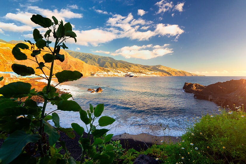 Ein malerischer Blick über eine kleine Bucht mit dunklem Sandstrand auf La Palma im warmen Licht des Spätnachmittags. Im Vordergrund sind grüne Pflanzen und gelbe Wildblumen zu sehen, die die Meeresbucht einrahmen. Das blaue Wasser spült sanft weiße Gischt an die Küste aus dunklem Lavgestein. Im Hintergrund erstrecken sich grüne Hänge und eine Küstenstadt vor den Bergen unter einem leicht bewölkten, blau-goldenen Himmel. La Palma Spanien Kueste
