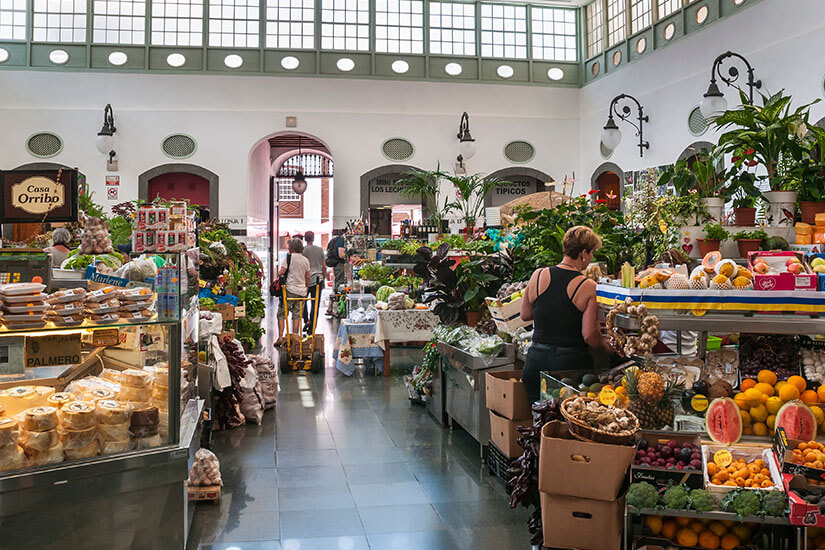 Ein Blick in eine belebte, lichtdurchflutete Markthalle mit hohen Decken und großen Fensterfronten. Auf der linken Seite präsentiert eine Theke frischen Käse und regionale Spezialitäten unter einem Schild mit der Aufschrift „Casa Orribo“. Auf der rechten Seite stehen reich gefüllte Stände mit frischem Obst und Gemüse wie Wassermelonen, Orangen, Papayas und Bananen sowie Zimmerpflanzen. Im Mittelgang bewegen sich Besucher zwischen den Marktständen. La Palma Spanien Markt