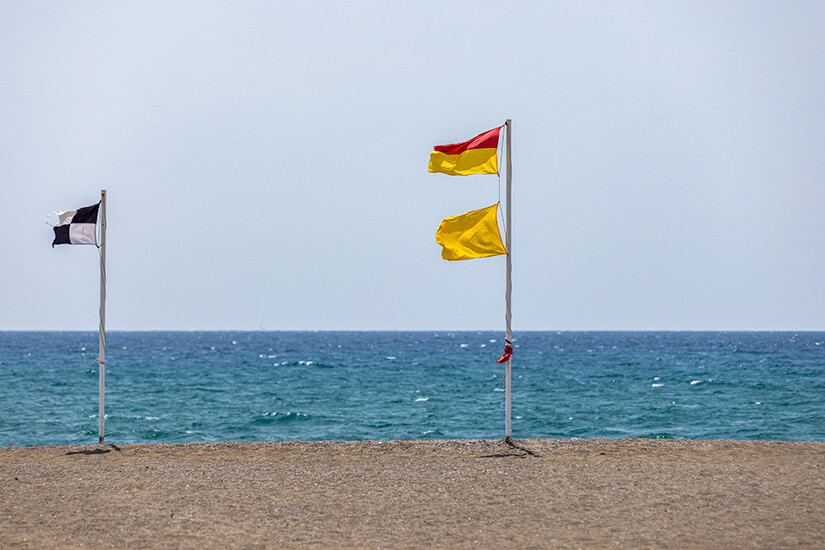 Eine schwarz-weiß karierte Flagge sowie eine gelbe und rotgelbe wehen am Strand im Wind. Flaggen am Strand bedeutung