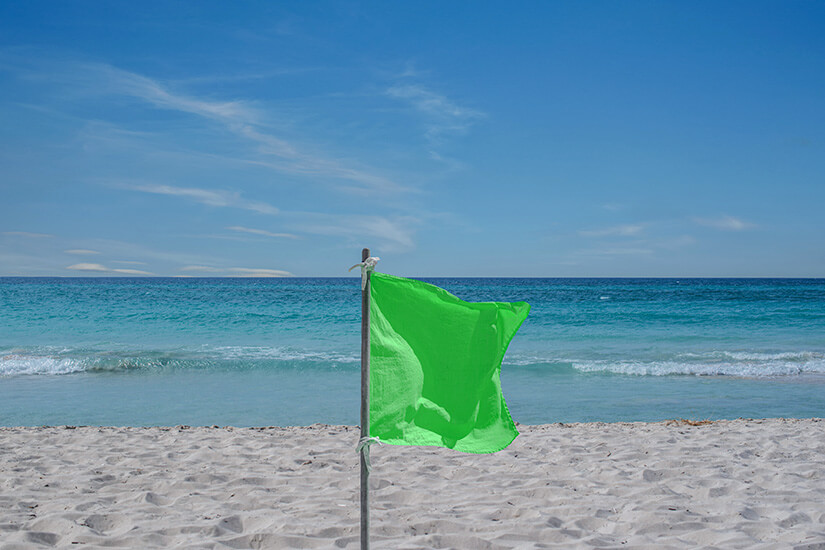 Die Nahaufnahme einer grünen Flagge an einem hellen Sandstrand. Ruhiges hellblaues Meer liegt im Hintergrund. Flaggen am Strand gruen