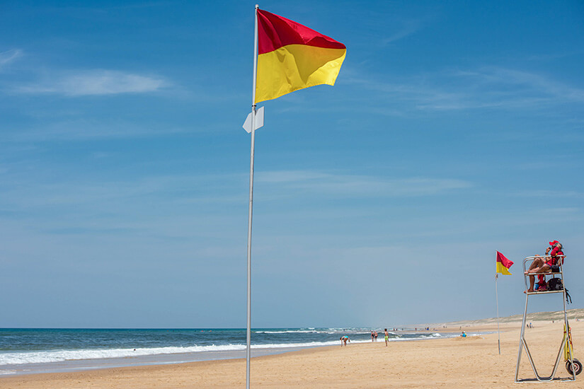 Zwei rot-gelbe Flaggen wehen am Strand. Ein Rettungsschwimmer sitzt auf einem Hochsitz, im Hintergrund sind Besucher zu sehen. Flaggen am Strand rot-gelb