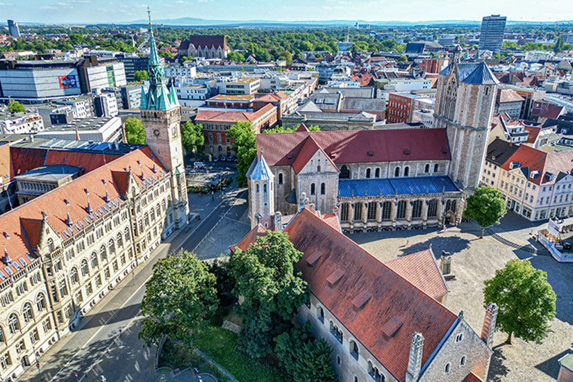 Zu erkennen ist eine Luftaufnahme einer historischen Innenstadt mit mehreren großen Kirchen, markanten Türmen und dicht bebauten Straßenzügen. Zwischen den historischen Bauwerken befinden sich moderne Gebäude, während sich am Horizont ausgedehnte Grünflächen und weitere Stadtteile erstrecken. Braunschweig Stadt