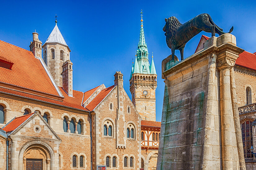 Zu erkennen ist eine historische Burganlage mit romanischen Gebäuden und mehreren markanten Türmen. Im Vordergrund steht ein großes Löwendenkmal auf einem steinernen Sockel, während der Himmel klar und tiefblau ist. Braunschweig Burg