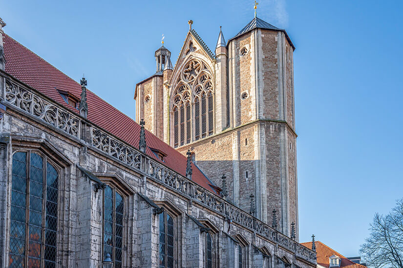 Zu erkennen ist eine imposante gotische Kirche mit hohen Steinmauern, einem markanten Turm und einem großen Maßwerkfenster. Im Vordergrund verläuft die reich verzierte Seitenfassade mit hohen Fenstern unter einem klaren blauen Himmel. Braunschweig Dom