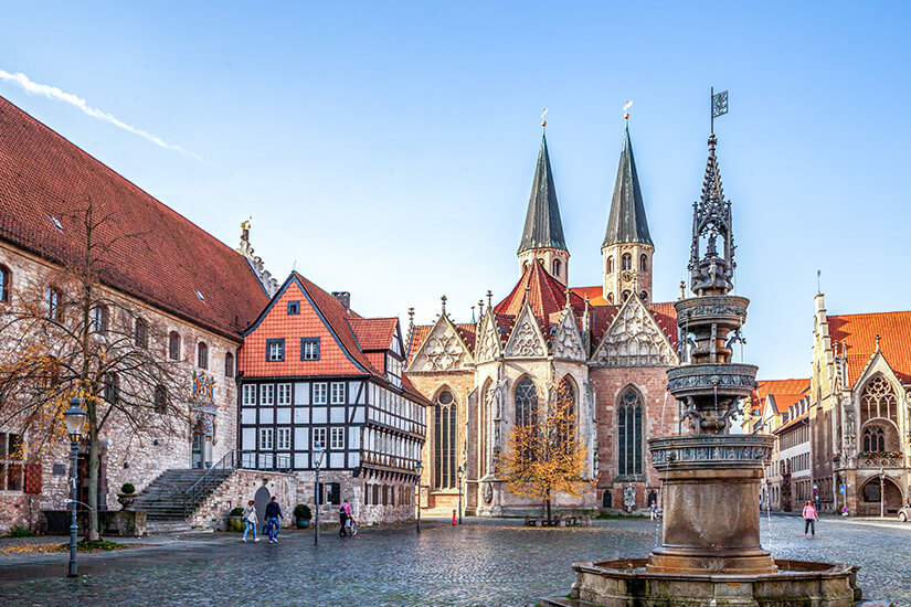 Zu erkennen ist ein historischer Platz mit einer gotischen Kirche, Fachwerkhäusern und weiteren historischen Gebäuden. Im Vordergrund steht ein kunstvoll verzierter Brunnen, während sich einige Menschen über das gepflasterte Areal bewegen. Braunschweig Altstadt