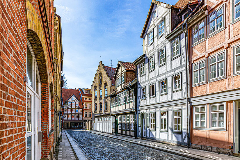 Zu erkennen ist eine schmale Kopfsteinpflasterstraße, die von gut erhaltenen Fachwerkhäusern und historischen Gebäuden gesäumt wird. Am Ende der Straße öffnet sich der Blick auf weitere Fachwerkbauten und ein markantes Gebäude mit gestuftem Giebel unter einem klaren blauen Himmel. Braunschweig Fachwerk