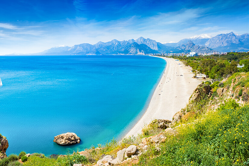Weit geschwungen erstreckt sich ein langer Kiesstrand entlang einer türkisblauen Bucht, im Hintergrund erheben sich beeindruckende schneebedeckte Bergketten unter strahlend blauem Himmel. Reiseziele August Tuerkei