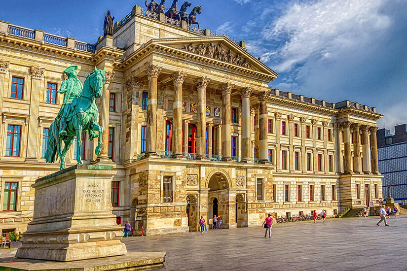 Zu erkennen ist ein repräsentatives Schloss oder Palais mit einer monumentalen Säulenfassade und einem großen Vorplatz. Im Vordergrund steht ein Reiterstandbild auf einem hohen Sockel, während sich auf dem Platz mehrere Menschen aufhalten. Braunschweig Schloss